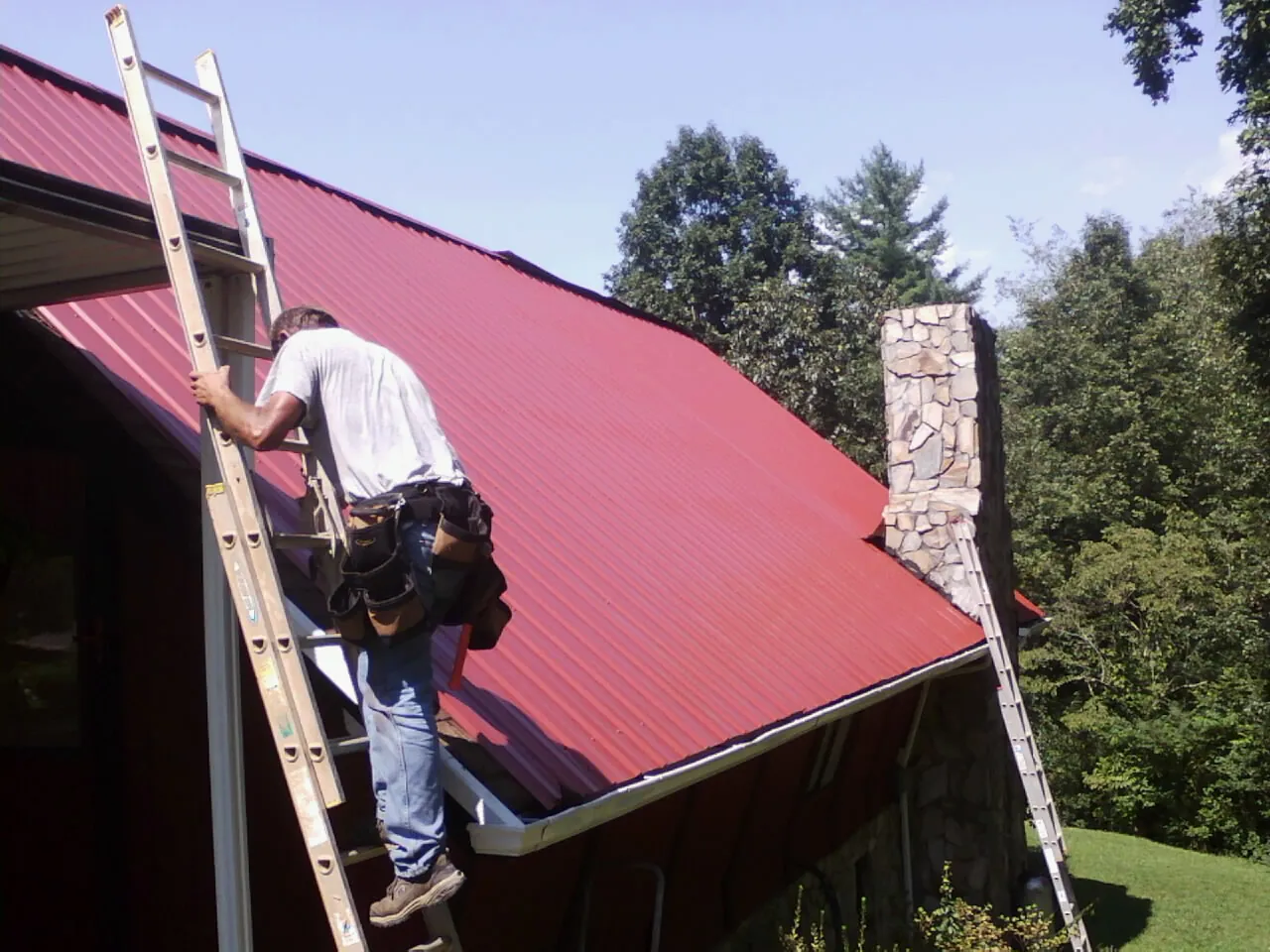 Roofer on a ladder beside a red metal roof and stone chimney — Soffit & Fascia Repair by Roofing Brothers in Romulus